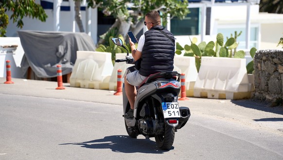 Mykonos, Greece - May 7, 2024: Young man uses his smartphone to make a call or video call on a scooter without a helmet. Danger on the road due to cell phone use Symbol photo *** Junger Mann benutzt s ...