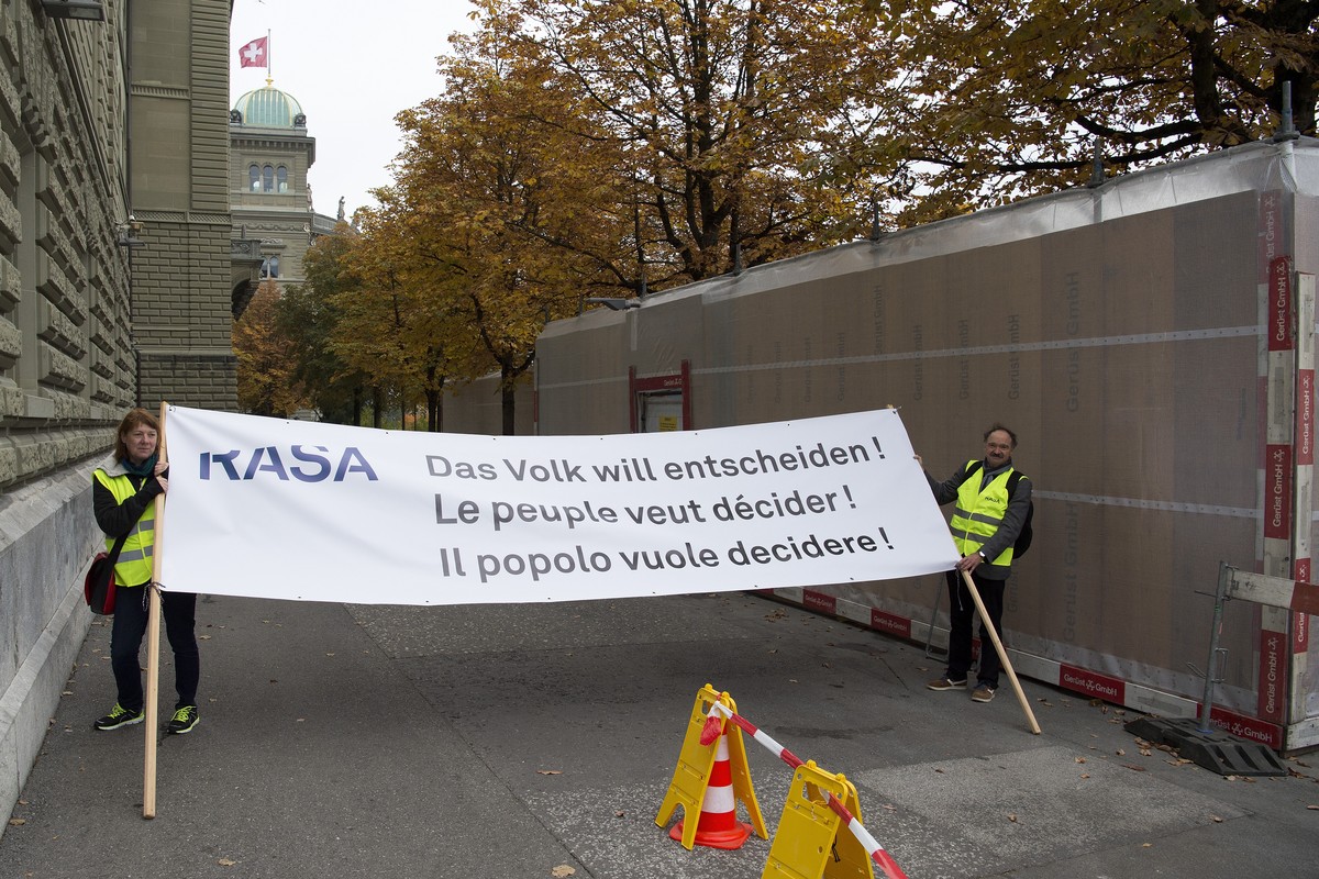 Zwie aktivisten halten ein Transparent mit der Aufschrift "Das Volk will entscheiden!, Le peuple veut decider!, Il popolo vuole decidere!", waehrend Aktivistinnen- und Aktivisten im Namen de ...