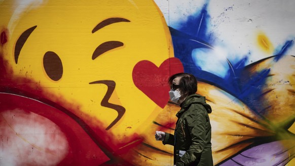 A woman wearing a protective face mask and gloves walks past a large emoji face painted on the boarded windows of a store in Vancouver, British Columbia, Wednesday, May 6, 2020. British Columbia has o ...