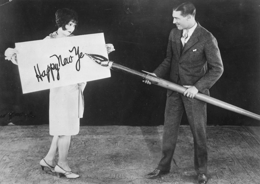 circa 1925: American actress Clara Bow (1905 - 1965) holds up a large card while actor Larry Gray inscribes a New Year&#039;s greeting with a giant pen. (Photo by General Photographic Agency/Getty Ima ...