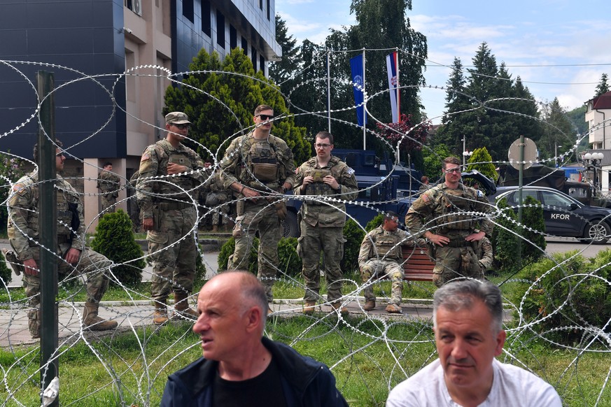 epa10668340 People sit in front of soldiers of the US contingent of the NATO-led international peacekeeping Kosovo Force (KFOR) while they take positions in front of the municipality building in Lepos ...