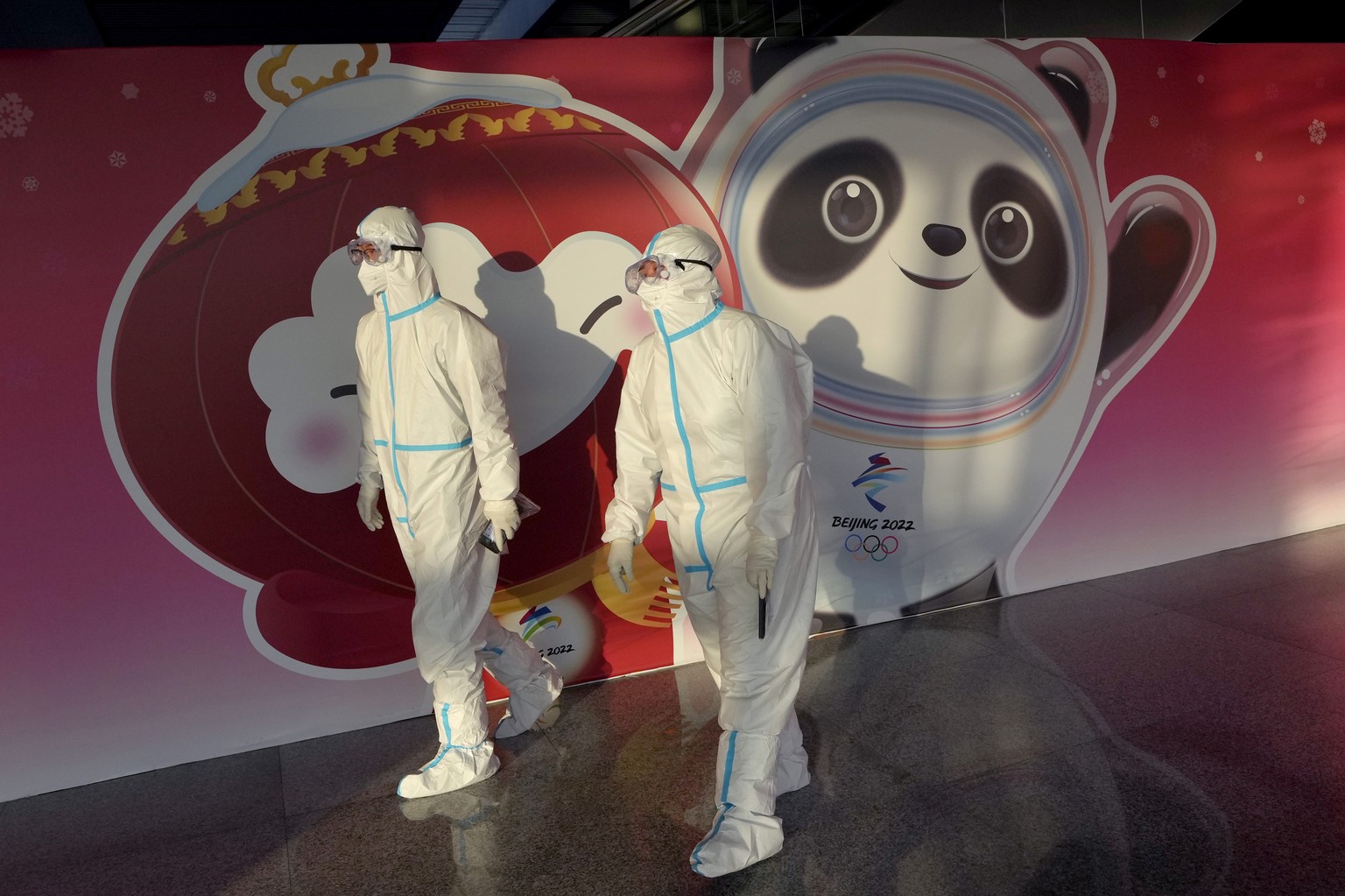 Olympic workers in protective gear walk through the Beijing Capital International Airport past a picture of Olympic mascot Bing Dwen Dwen, and Paralympic mascot Shuey Rhon Rhon, left, as they work to  ...