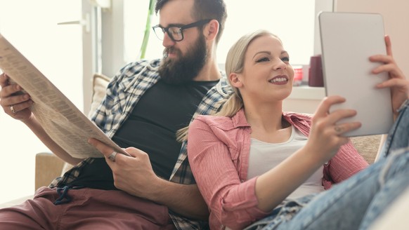 Junges verliebtes Paar sitzt auf einer Couch in ihrer Wohnung neben dem Fenster, genießen ihre freie Zeit, Zeitung lesen und im Internet surfen auf einem Tablet-Computer. Lens Flare Effekt auf Fenster