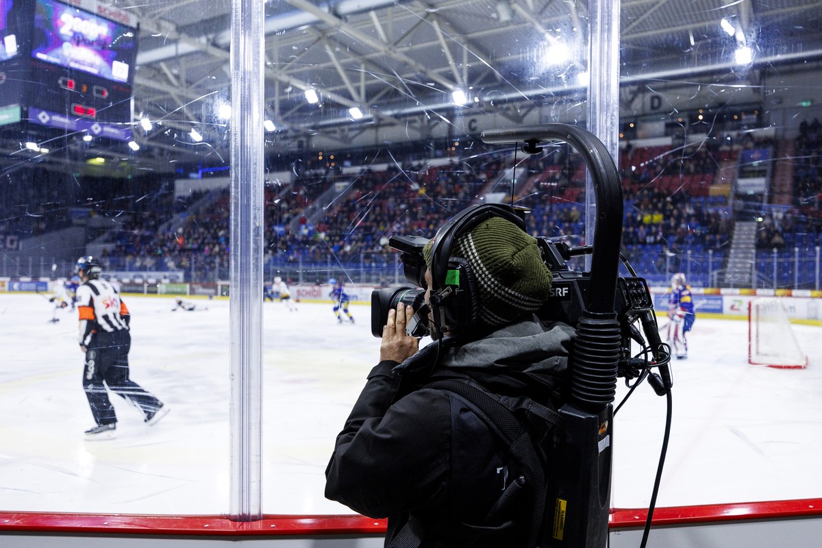 Ein SRF TV Kameramann filmt durch die Plexiglas-Scheibe das Eishockey-Meisterschaftsspiel der National League zwischen den Teams EHC Kloten und HC Ajoie am Samstag, 2. Dezember 2023, in Kloten. (KEYST ...