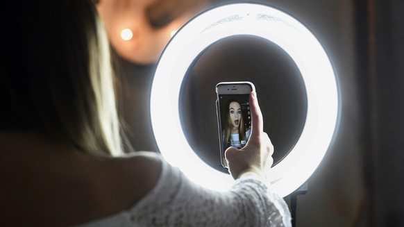In this Feb. 28, 2018 photo, Matty Nev Luby holds up her phone in front of a ring light she uses to lip-sync with the smartphone app Musical.ly, in Wethersfield, Conn. Teens and young adults say cyber ...