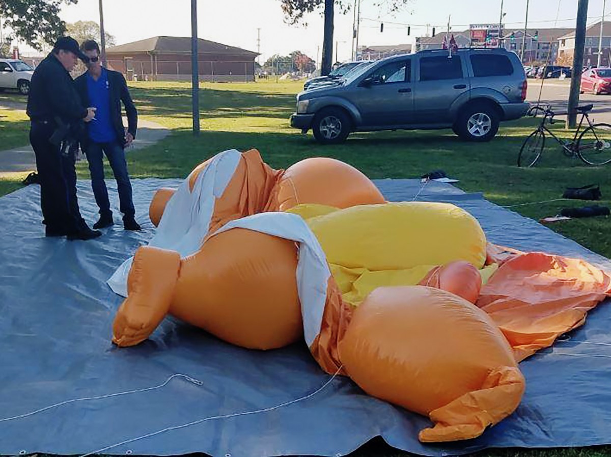 A responding officer and an unidentified man stands by a Baby Trump balloon deflated by someone at Monnish Park as people were protesting President Donald Trump's visit to an NCAA college footbal ...