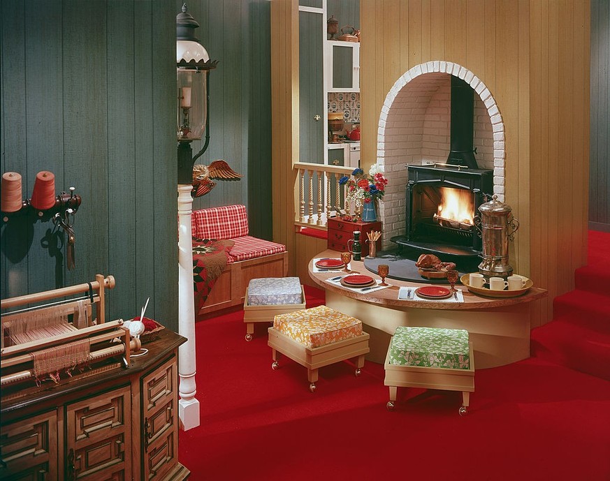 1970: A model dining room features a wood stove in a brick-lined arched nook, a table with three place settings, a samovar and mugs, a credenza with a small loom, a lamppost, three stools on casters,  ...