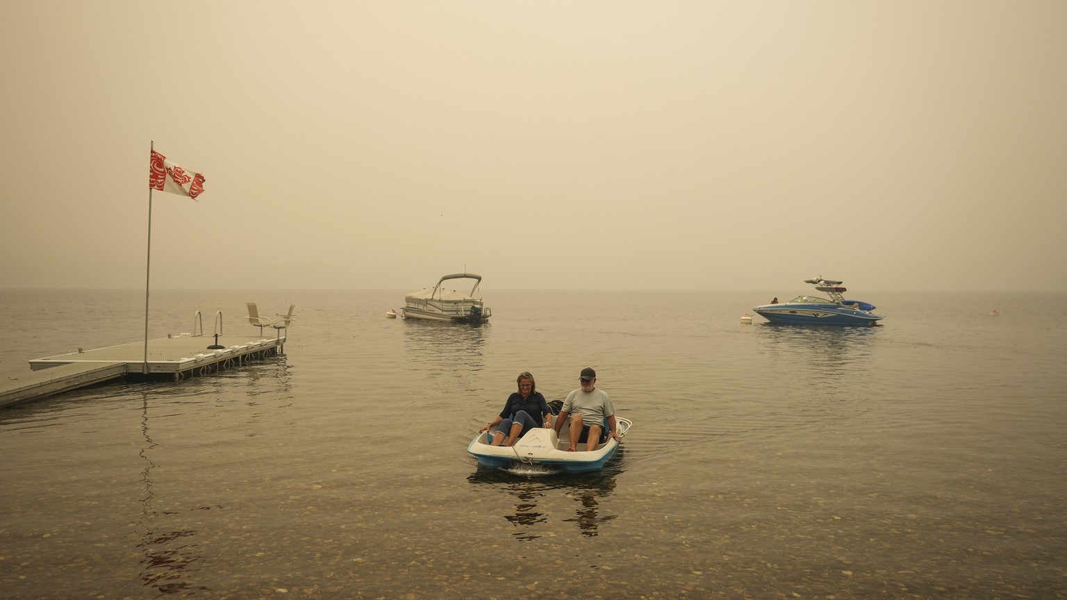 Smoke fills the air as Pat Manzuik and his wife Trevor use a paddleboat to get to shore after being given a boat ride by good samaritan Christy Dewalt, back right, back to their home they were evacuat ...