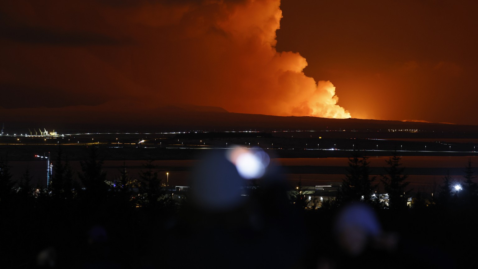 People watch as the night sky is illuminated caused by the eruption of a volcano on the Reykjanes peninsula of south-west Iceland seen from the capital city of Reykjavik, Monday Dec. 18, 2023. (AP Pho ...