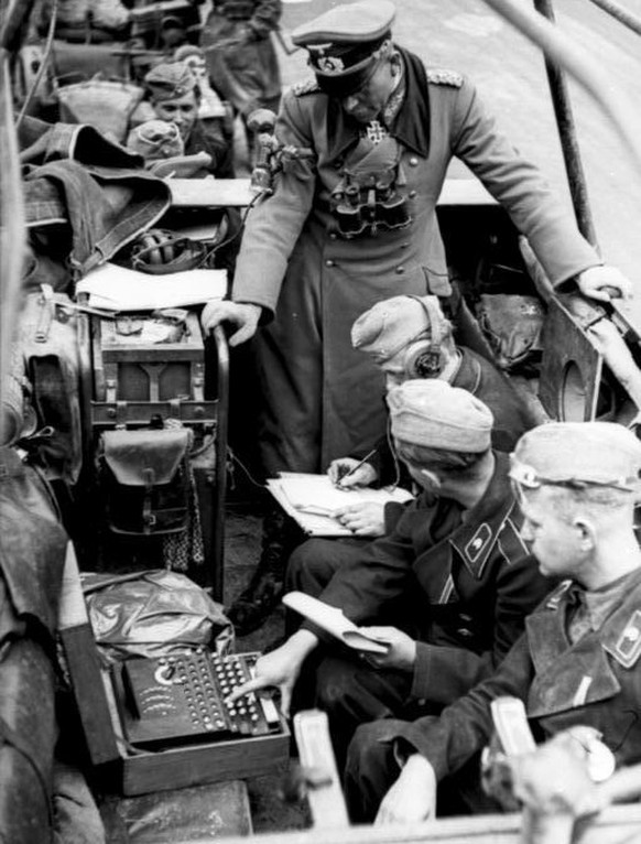 Eine Enigma im Einsatz während des Zweiten Weltkriegs. Unter den Augen von General Heinz Guderian empfängt ein Funker eine Nachricht, die von einem Soldaten an der Enigma entschlüsselt wird. Auf der Tastatur wird die verschlüsselte Nachricht Buchstabe für Buchstabe eingegeben, im Bereich darüber leuchten die jeweils entschlüsselten Buchstaben auf.