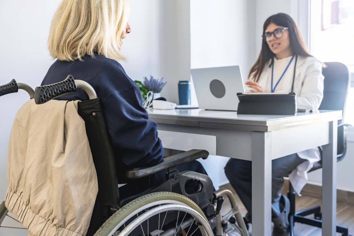 Patientin im Rollstuhl, die während einer medizinischen Beratung in einer hellen Klinikumgebung ärztlichen Rat von einer Ärztin erhält, wobei der Schwerpunkt auf der Betreuung und Unterstützung von Be ...