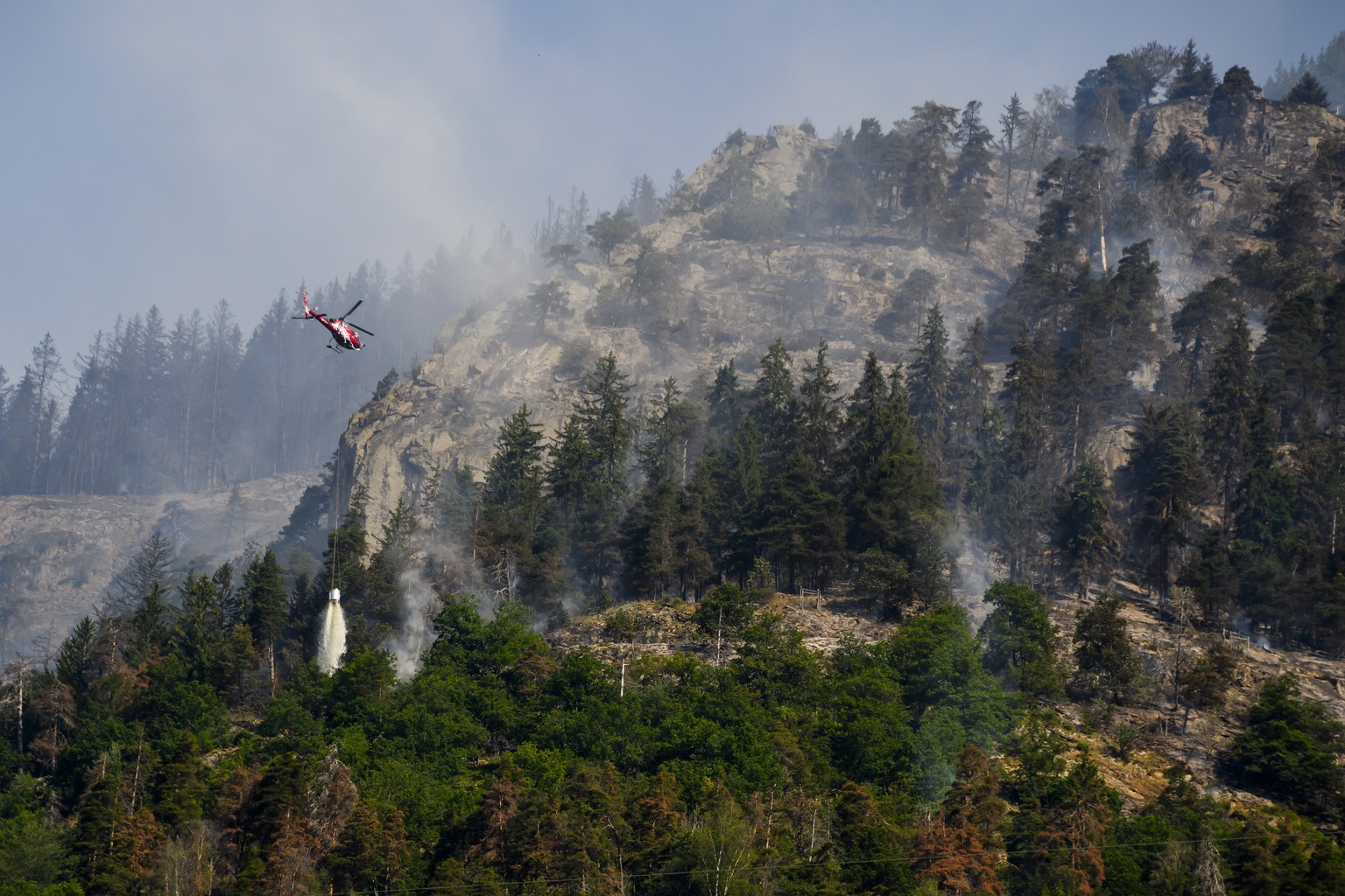 epa10753042 A helicopter pours water on the still burning forest above the communes of Bitsch and Ried-Moerel, in Bitsch, Switzerland, 18 July 2023. A forest fire broke out above Bitsch/Ried-Moerel in ...