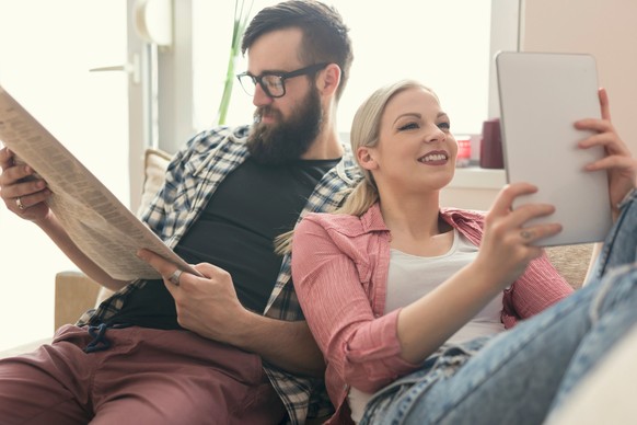 Junges verliebtes Paar sitzt auf einer Couch in ihrer Wohnung neben dem Fenster, genießen ihre freie Zeit, Zeitung lesen und im Internet surfen auf einem Tablet-Computer. Lens Flare Effekt auf Fenster