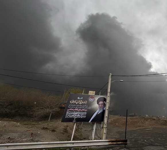 A thick plume of smoke from a U.S.-Israeli strike on an oil facility late Saturday lingers in the cloudy sky behind a billboard showing the late Iranian Supreme Leader Ali Khamenei in Tehran, Iran, Su ...