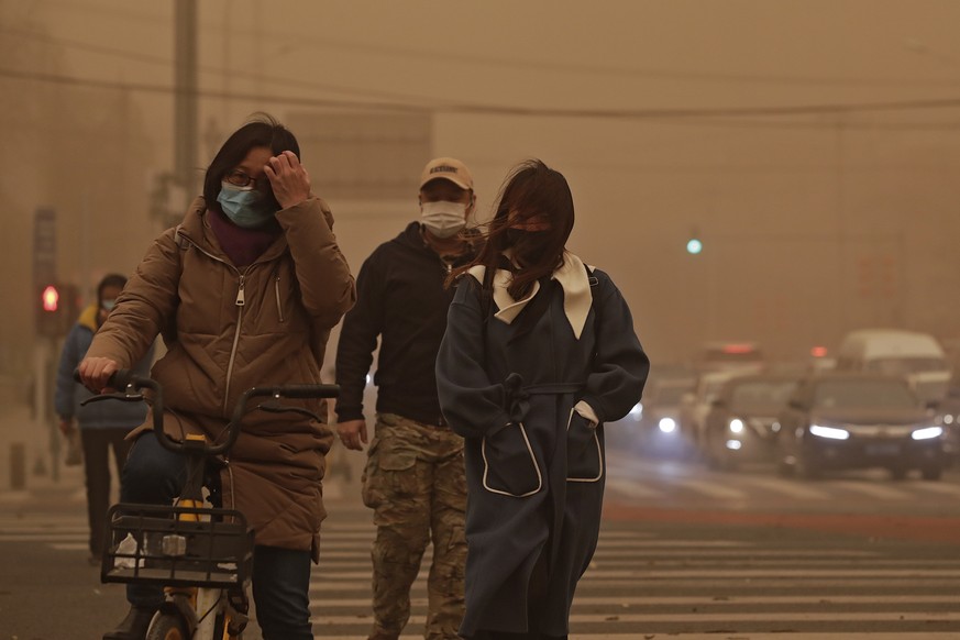 People wearing face masks walk across a street as capital city is hit by polluted air and sandstorm in Beijing, Monday, March 15, 2021. The sandstorm brought a tinted haze to Beijing's skies and ...