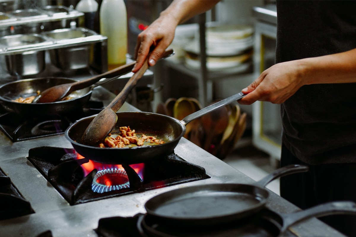 eine Person, die in einer professionellen Küche kocht, Zutaten in einer Pfanne über einer Gasflamme rühren. Der Koch verwendet einen Holzlöffel und einen Metallspatel, um anscheinend Fleischstücke ode ...