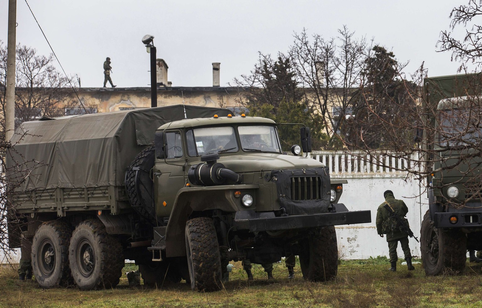 Bewaffnete Männer, vermutlich russische Soldaten, patrouillieren vor einer ukrainischen Militärbasis in Sewastopol. Auf dem Dach im Hintergrund ein ukrainischer Soldat.&nbsp;