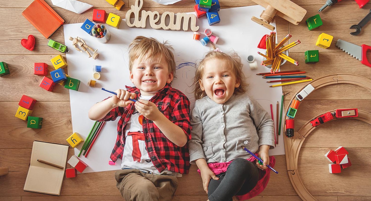 Kids drawing on floor on paper. Preschool boy and girl play on floor with educational toys - blocks, train, railroad, plane. Toys for preschool and kindergarten. Children at home or daycare. Top view