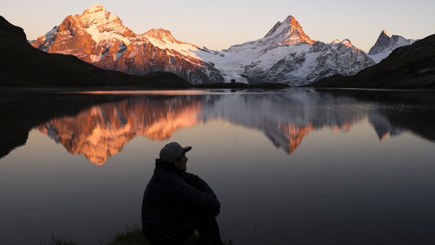 A man looks at the mountains Wetterhorn, Lauteraarhorn, Schreckhorn and Finsteraarhorn that are reflecting during the sunset in the Bachalpsee lake (2265m) close to the First, above Grindelwald in the ...
