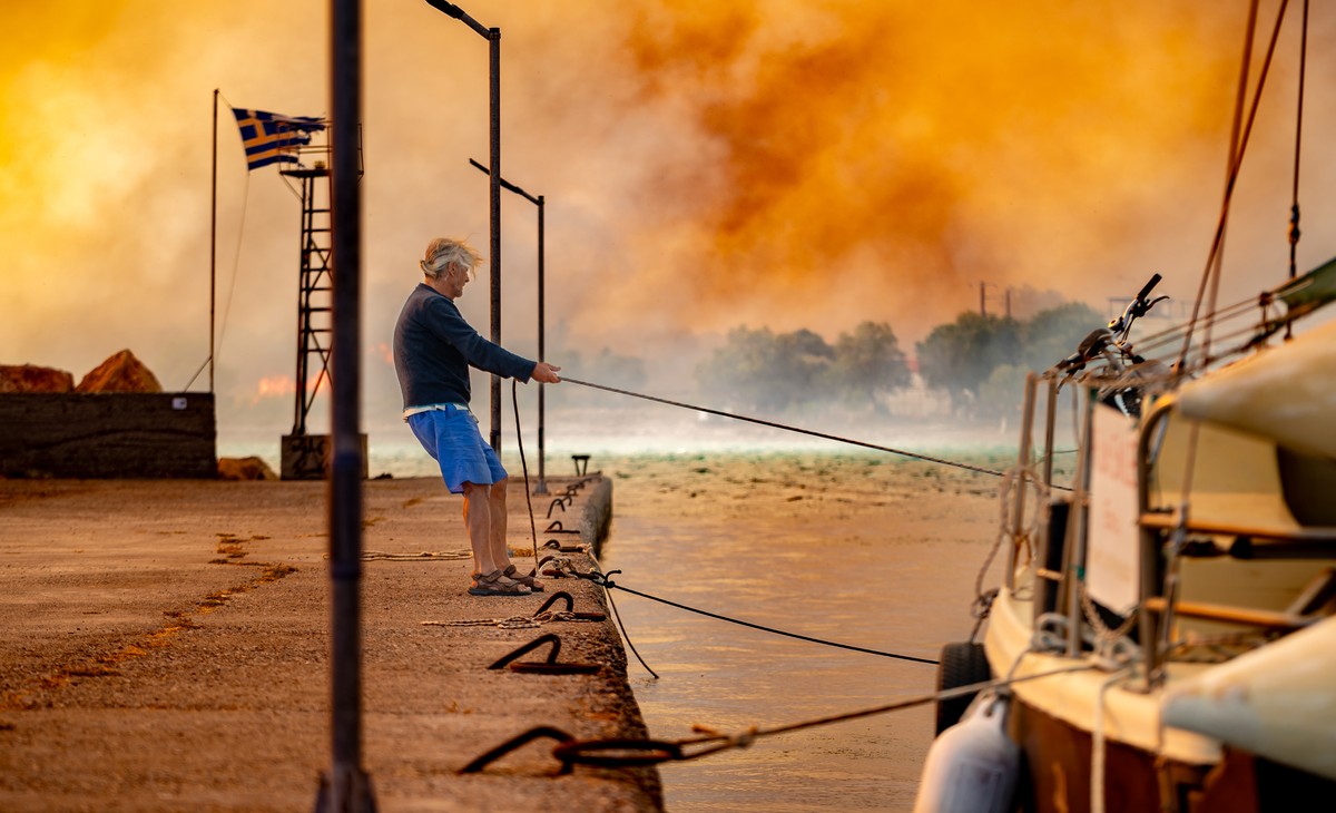 KEYPIX - epa12196792 A man ties a boat at the port of Lithi in Chios, while a wildfire burns through forested land, Greece, 24 June 2025 (issued 25 June 2025). Greek authorities on 25 June announced t ...