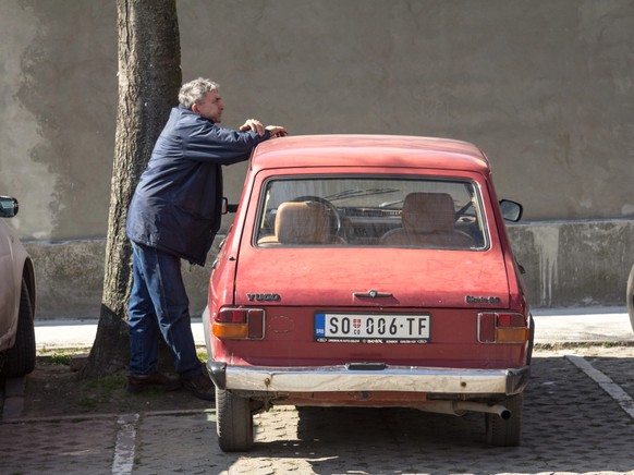 SOMBOR, SERBIA - MARCH 19, 2016: Senior old man, serb, standing on a Zastava and Yugo 55 red car. Also known as Skala, it&#039;s a family of cars built by Serbian manufacturer Zastava Automobili, base ...