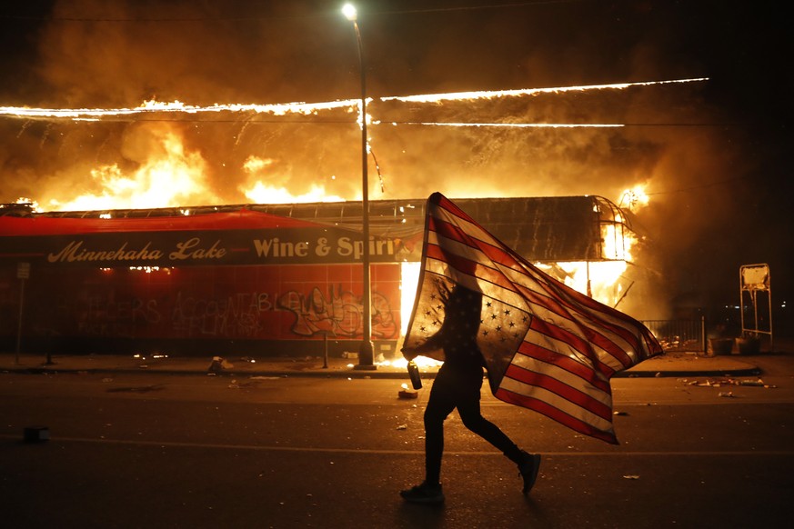 A protester carries a U.S. flag upside down, a sign of distress, next to a burning building Thursday, May 28, 2020, in Minneapolis. Protests over the death of George Floyd, a black man who died in pol ...