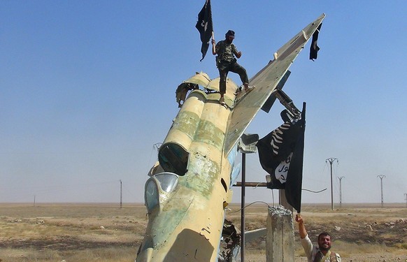 Kämpfer der Terrorgruppe IS posieren mit ihrer schwarzen Flagge vor einem zerstörten syrischen Kampfflugzeug (Archivbild).