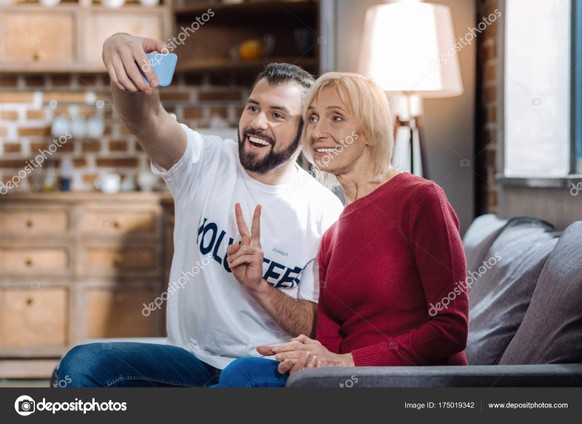 Cool photo. Cheerful funny positive social worker sitting on the sofa with a senior woman and taking photos with her