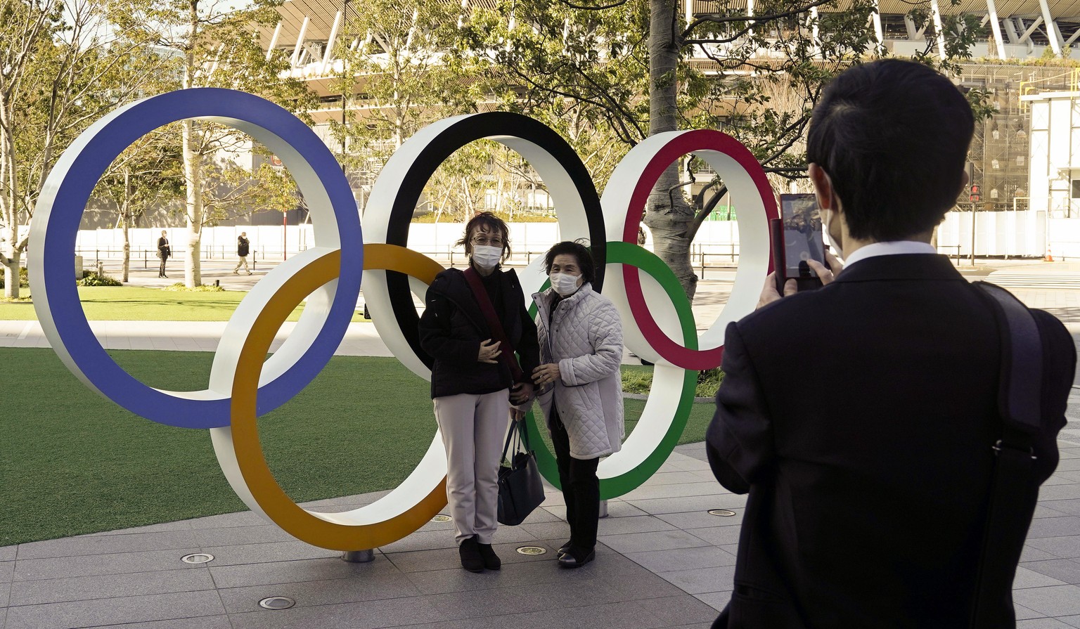 epa08251625 Visitors wearing face masks take a commemorating photo at the Olympic Rings monument in front of the Japan Olympic Committee headquarters near the new National Stadium (back), venue of the ...