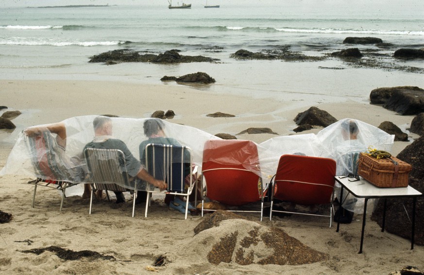 People, Holidays, England, pic: 1970, Hardy holidaymakers hoping for a picnic have to endure the British climate as they take shelter on the beach by covering themselves in a plastic sheet (Photo by B ...