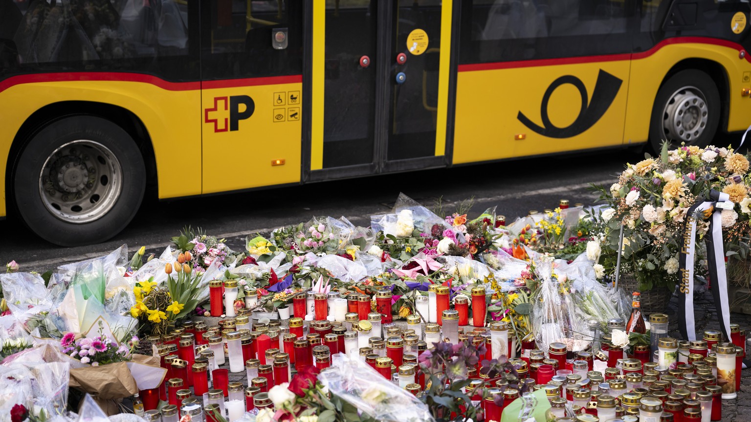 KEYPIX - Ein Postauto faehrt an Blumen und Kerzen vorbei, an der Stelle, an der ein Postauto in Brand geriet, am Sonntag, 15. Maerz 2026, im Zentrum von Kerzers im Kanton Freiburg. Laut der Freiburger ...