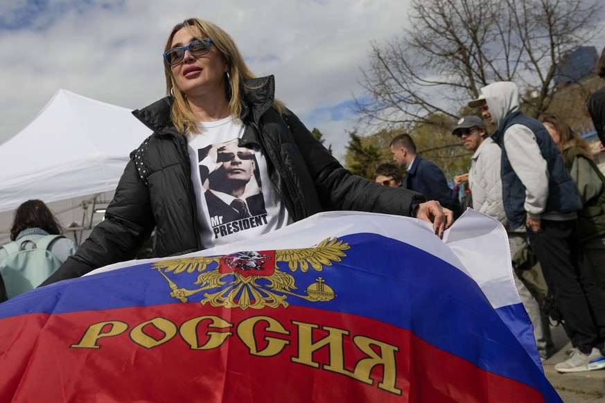 A woman wearing a shirt with a picture of Russian President Vladimir Putin holds a Russian flag outside a polling station set up in a Russian school in Belgrade, Serbia, Sunday, March 17, 2024. Russia ...
