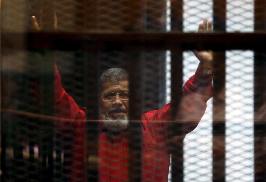 Egypt&#039;s deposed president Mohamed Mursi greets his lawyers and people from behind bars at a court wearing the red uniform of a prisoner sentenced to death, during his court appearance with Muslim ...