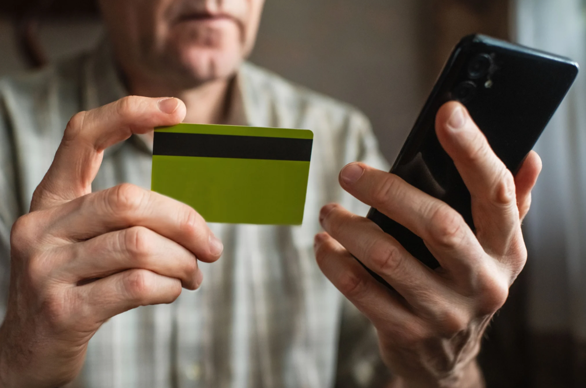 Ein Senior mit Smartphone hält eine Bankkarte in seinen Händen. Echte Bankangestellte würden nie verlangen, die Karte zu zerschneiden und sie einer Drittperson zu übergeben.