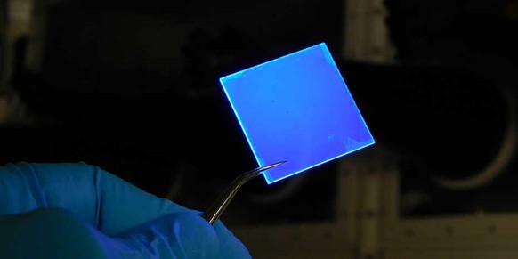 Eine mit mehreren Lagen extrem dünner Halbleiter-Nanoplättchen beschichtete Glasscheibe, die mit UV-Licht beschienen wird und blaues Licht aussendet.