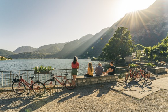 Eine Freundesgruppe unternimmt die Velotour mit Guide und macht eine Pause am Seeufer, während die Sonne über dem See aufgeht.
