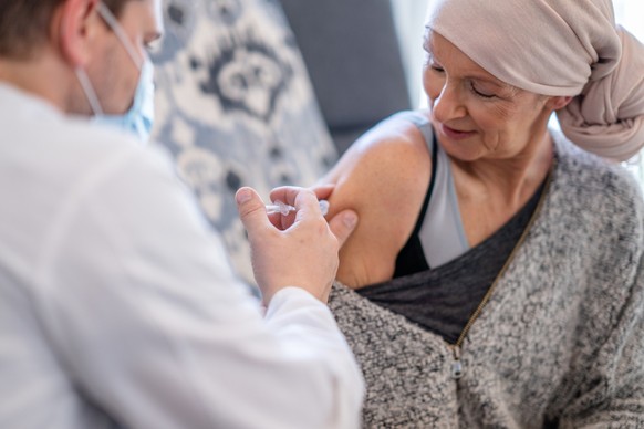 A older woman wearing a head scarfs smiles in a relaxed manner as her visiting doctor administers an injection to her in her shoulder. Krebs Impfung vaccine cancer Spritze Symbolbild