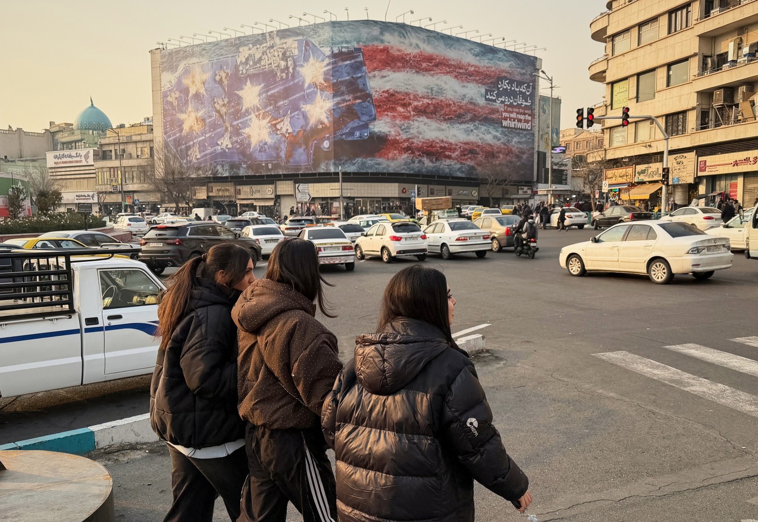 People walk in front a billboard with graphic showing a U.S aircraft carrier with damaged fighter jets on its deck, and sign reading in Farsi and English: "If you sow the wind, you'll reap w ...