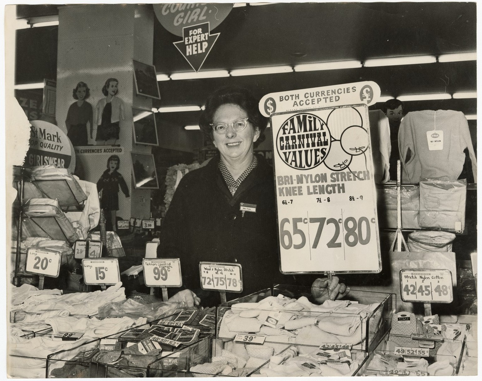 Mrs A. Tipa, a member of staff at Woolworths stood in front of a display of socks. Prices are displayed in pounds and dollars due to changeover to decimal currency.