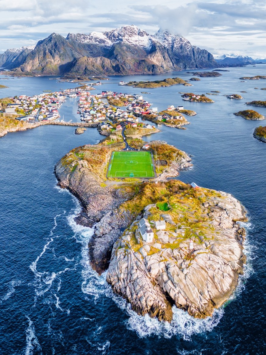 A stunning aerial view of Henningsvær in Norway’s Lofoten Islands shows colorful houses, dramatic mountains, scattered islets, and the iconic seaside football field.