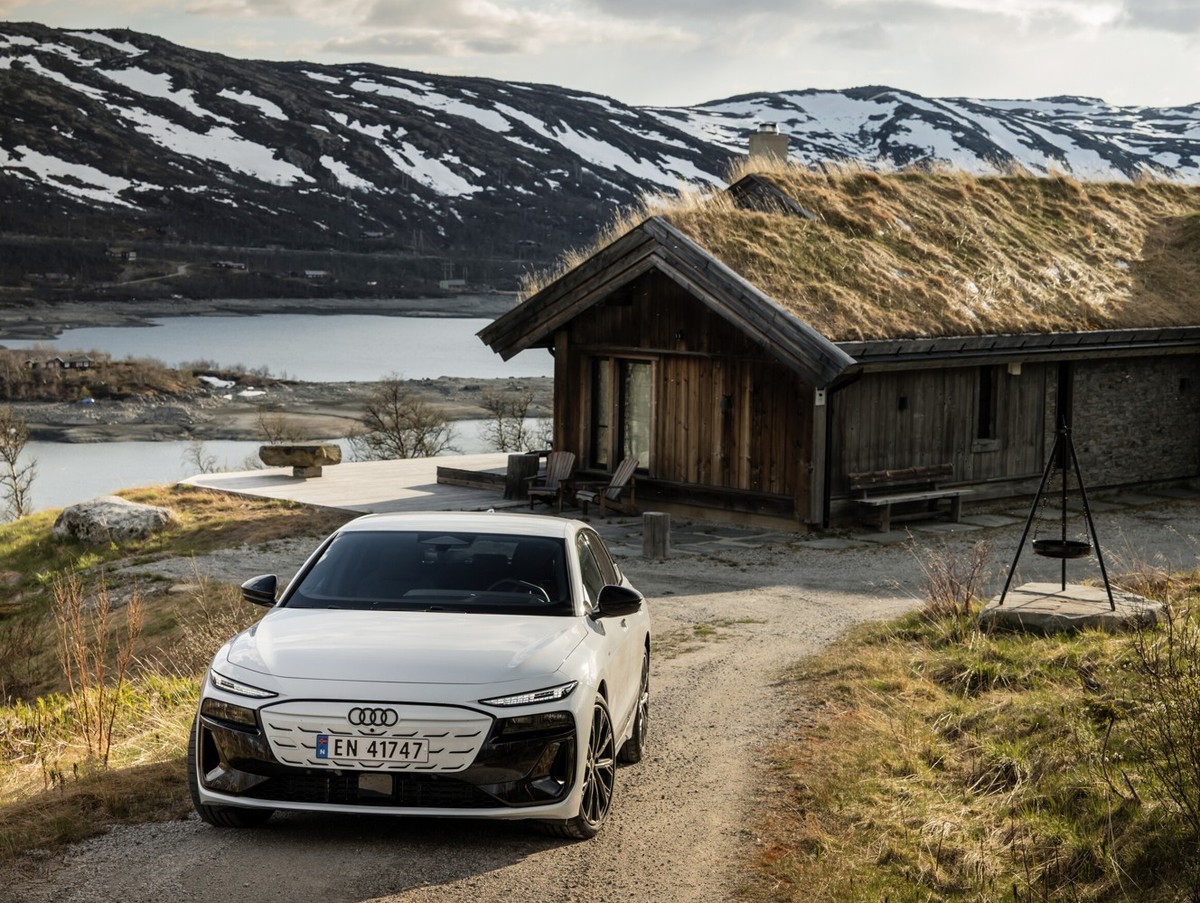 An Audi A6 Sportback e-tron in Glacier white metallic is parked on a gravel road that leads to a house of classic Scandinavian design.