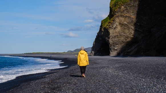 Ein Spaziergang am malerischen Reynisfjara-Strand in Island an einem sonnigen Tag A traveler enjoys a peaceful walk on Reynisfjara Beach in Iceland, known for its black sand and stunning cliffs. The s ...