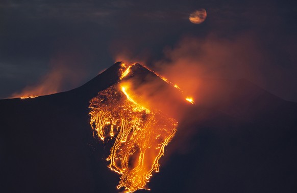 The moon is partially seen in the sky as lava flows from the Mt. Etna volcano, near Catania in Sicily, southern Italy, early Tuesday, Feb. 23 , 2021. An explosion started Monday evening, provoking a h ...