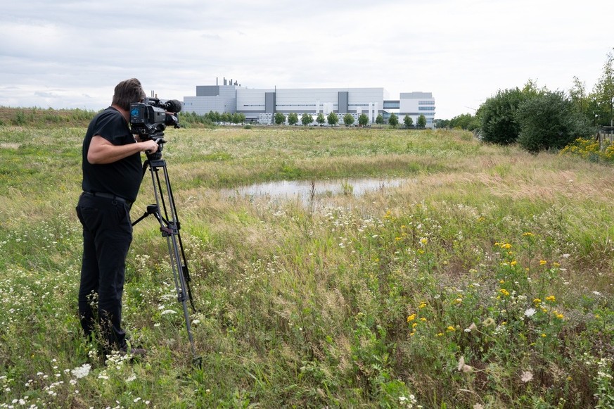 08.08.2023, Sachsen, Dresden: Ein Kameramann steht auf einer Wiese in einem Industriepark im Dresdner Stadtteil Klotzsche. Im Hintergrund ist die Halbleiterfabrik von Bosch zu sehen. Der taiwanische C ...