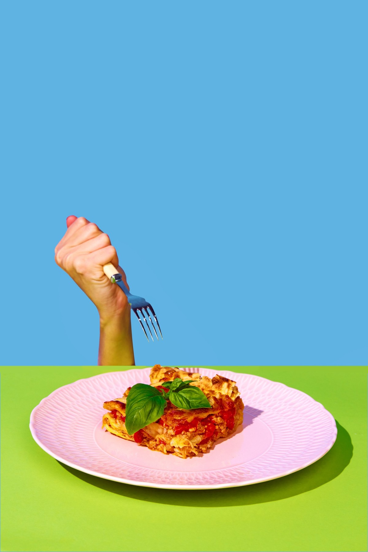 Female hand holding fork over delicious Italian food, lasagna over green and blue background. Bolognese sauce and basil. Concept of Italian food, cuisine, taste, cooking, menu. Pop art. Poster, ad