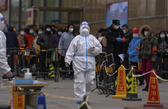 A health worker wearing a protective suit walks by masked residents who wait in line to get their throat swab at a coronavirus testing site following a COVID-19 case was detected in a residential buil ...