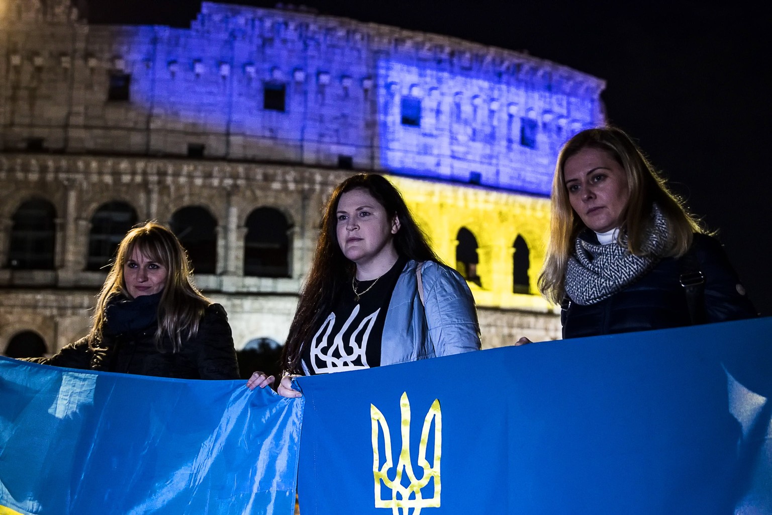 epa09782356 People hold Ukrainian flags as the Colosseum is illuminated with the colors of the Ukrainian flag in solidarity with the Ukrainian people, in Rome, Italy, 24 February 2022. Russian troops  ...