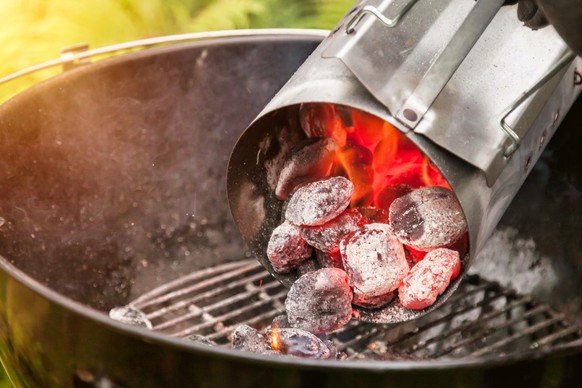Glowing red-hot charcoal is being poured from a metal starter chimney into a round grill. The coals are landing on the grill rack, ready for cooking.