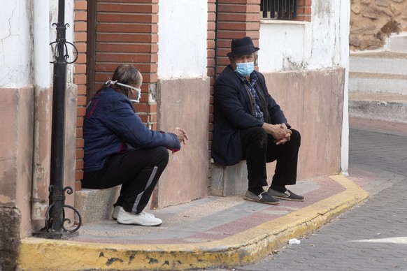 Two men wearing face masks to protect against the spread of coronavirus sit and talk on a street corner in the rural village of La Puerta de Segura, Jaen, Spain, Tuesday, Oct. 27, 2020. Despite overnight curfews, mobility curbs affecting at least four regions and other restrictions, health authorities in Spain are warning that sharp outbreaks of Covid-19 are already straining hospitals. (AP Photo/Paul White)