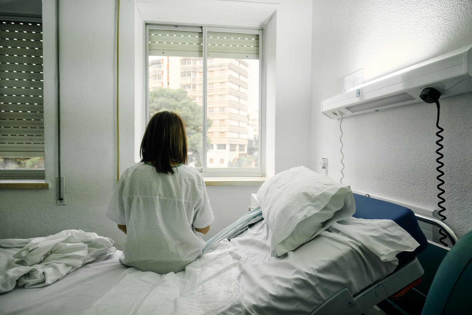 sick woman alone sitting on the bed of a hospital room looking out the window seen from behind